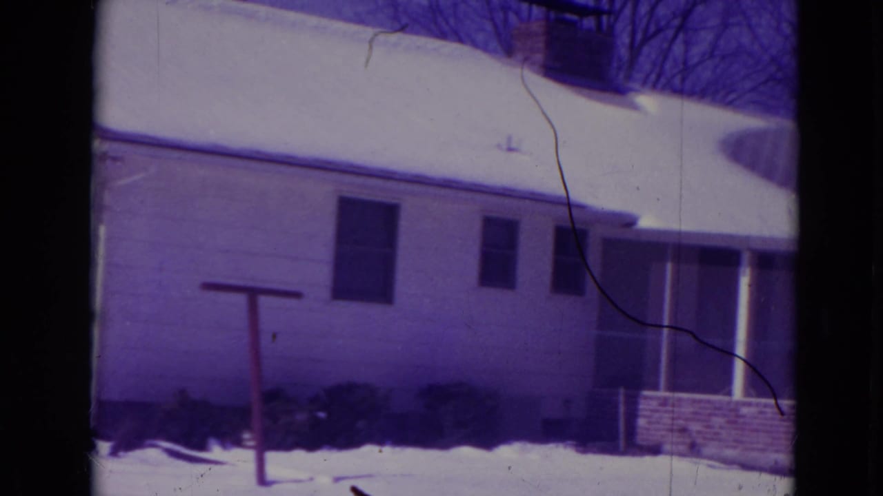 Side of a snow-covered house with shrubs, windows, and a small yard post.