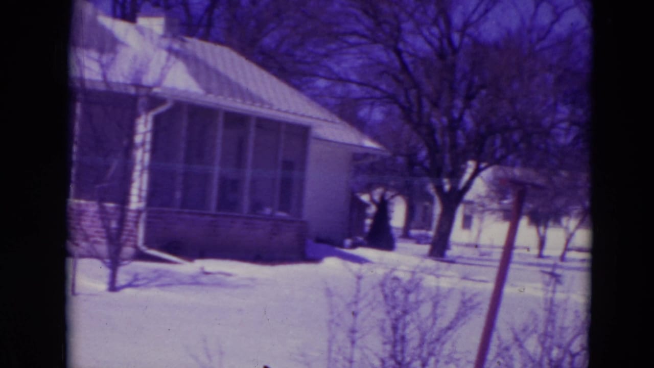 Snow-covered yard beside a one-story house with bare trees and a residential street beyond.