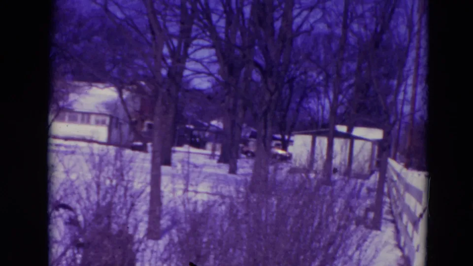 1974 8mm Orlando frame showing a snowy backyard or side yard with trees, shrubs, a shed, fencing, and distant cars.