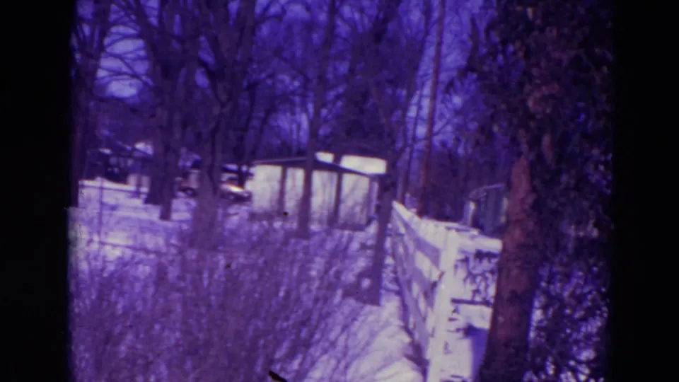 Final 1974 Orlando frame showing a snow-covered yard, small shed, fence, bare trees, shrubs, and distant houses or vehicles.