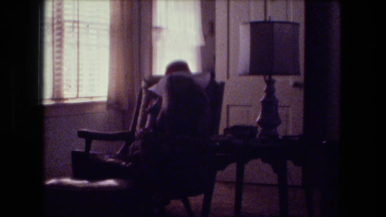 1974 Fort Wayne home-movie frame showing a seated figure in a wooden armchair near window blinds, curtains, and a table lamp.