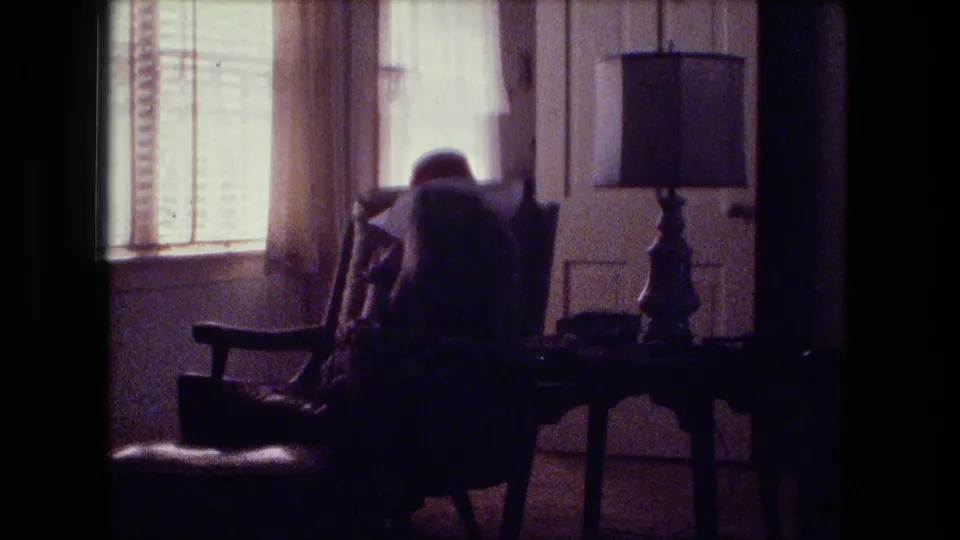 1974 Fort Wayne home-movie frame showing a seated figure in a wooden armchair near window blinds, curtains, and a table lamp.
