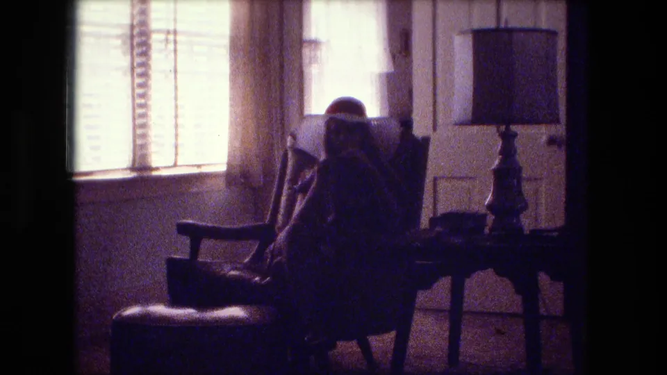 A grainy 8mm frame shows a seated figure in a Fort Wayne living room near curtains, blinds, a lamp, and paneled doors.