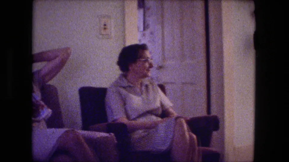 In a Fort Wayne living room, a woman in glasses sits in a chair while another seated person is partly visible at the edge of the frame.