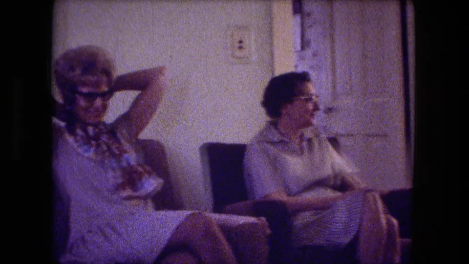 Fort Wayne 1974 home-movie frame showing two women seated in a living room, with one leaning back and adjusting her hair.
