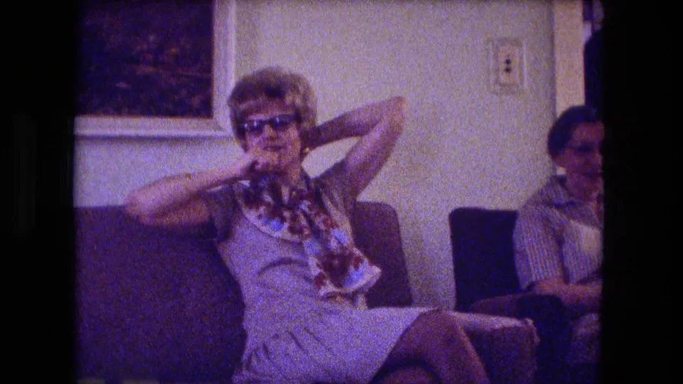 Fort Wayne 1974 home interior frame showing a woman in sunglasses adjusting her scarf or hair on a couch.