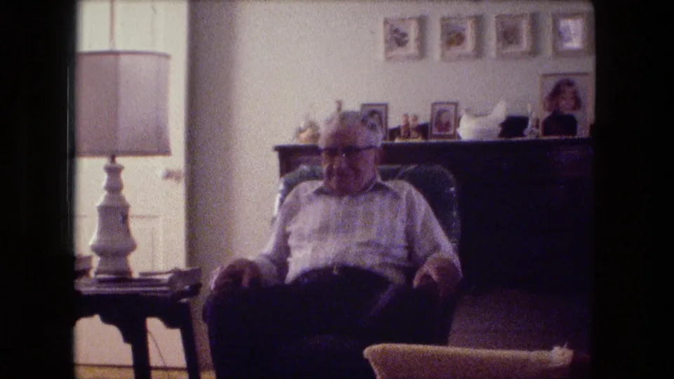 Fort Wayne 1974 living-room frame showing an older man seated in an armchair beside a lamp and mantel photographs.