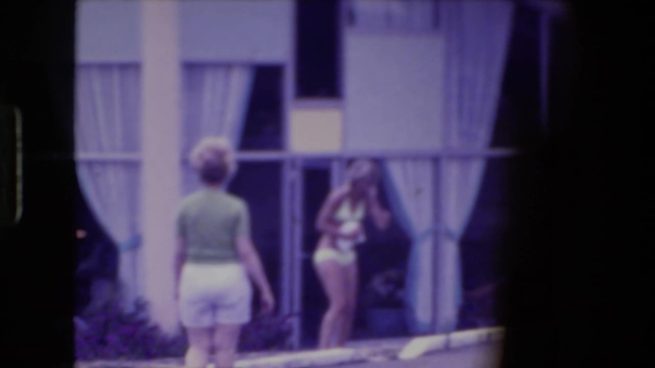 1974 Fort Wayne home-movie frame showing two women in summer clothing near a poolside building with sheer curtains.