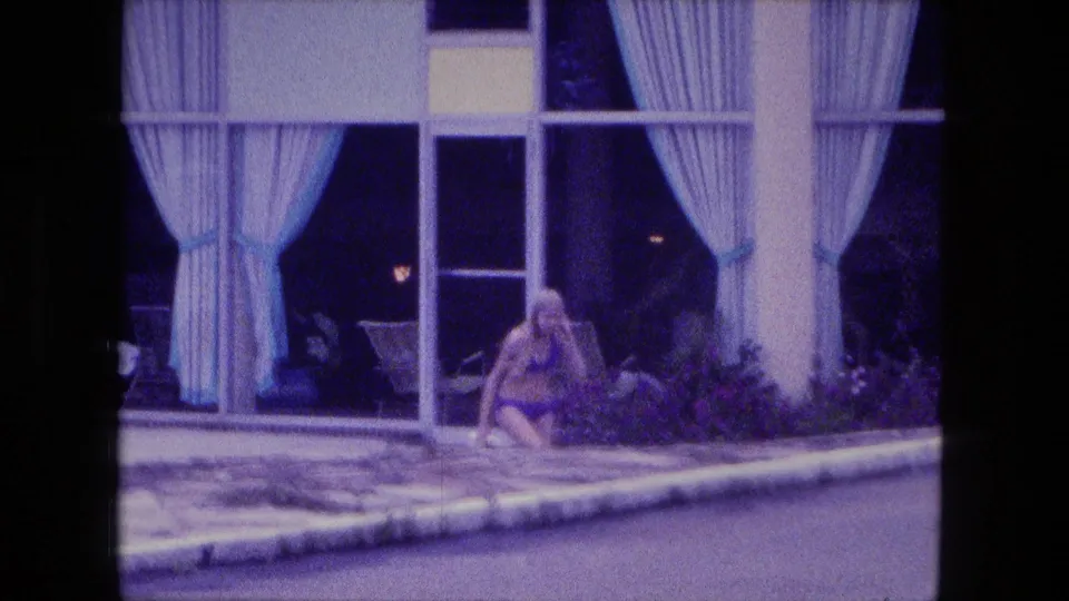 1974 Fort Wayne poolside frame with a woman in a bikini low near the walkway in front of curtained windows.