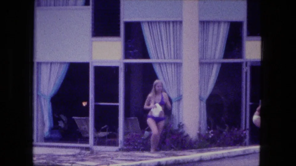 Fort Wayne 1974 home-movie frame showing a woman in a bikini carrying a towel near a curtained poolside entrance.