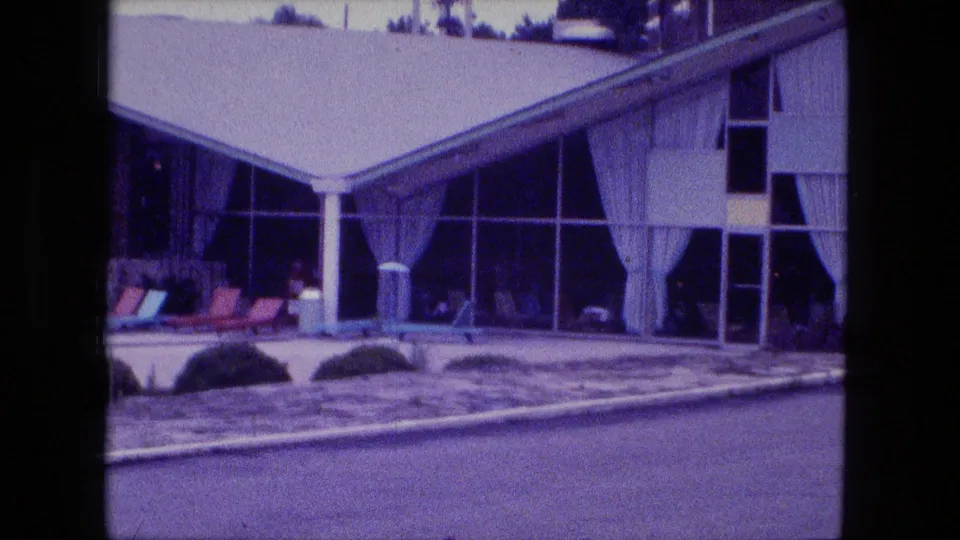 1974 Fort Wayne 8mm frame showing lounge chairs beside a pool and the curtained glass facade of a sloped-roof modernist building.