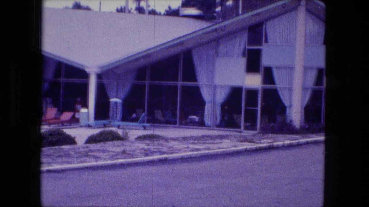 1974 Fort Wayne motel exterior showing a modernist glass entrance, sloped roof, white curtains, patio seating, and landscaped grounds.