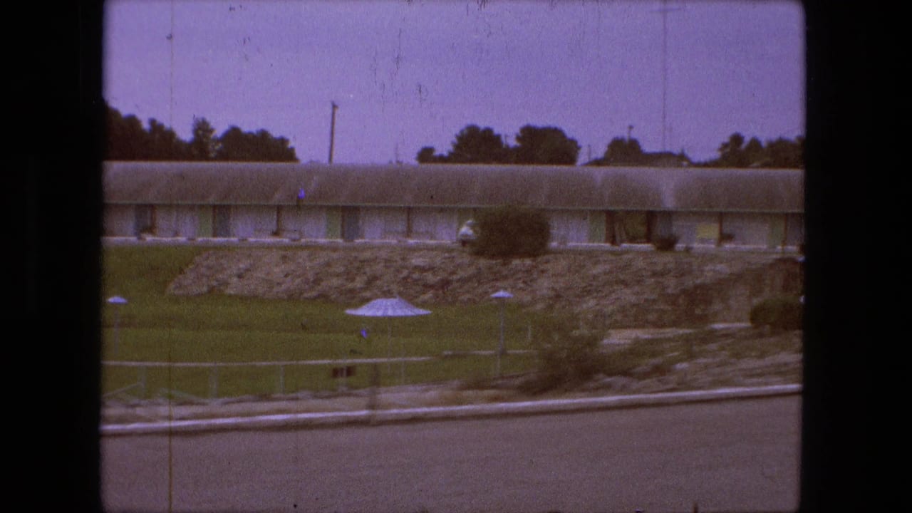Long motel room wing behind a lawn, stone landscaping, shrubs, and small umbrella-like fixtures.