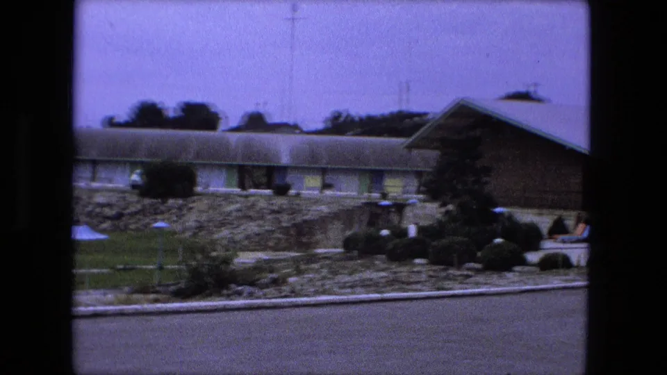 1974 motel exterior in Fort Wayne showing a long room wing, brick side wall, landscaped rocks, shrubs, and patio lounge chairs.