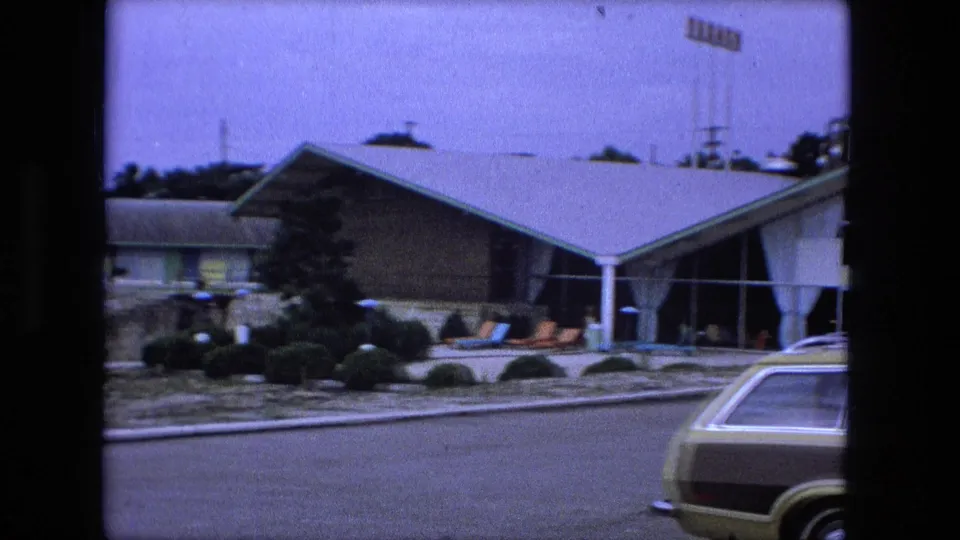 Fort Wayne motel exterior in 1974 with sloped-roof glass entrance, patio chairs, landscaped shrubs, road, and a classic station wagon.