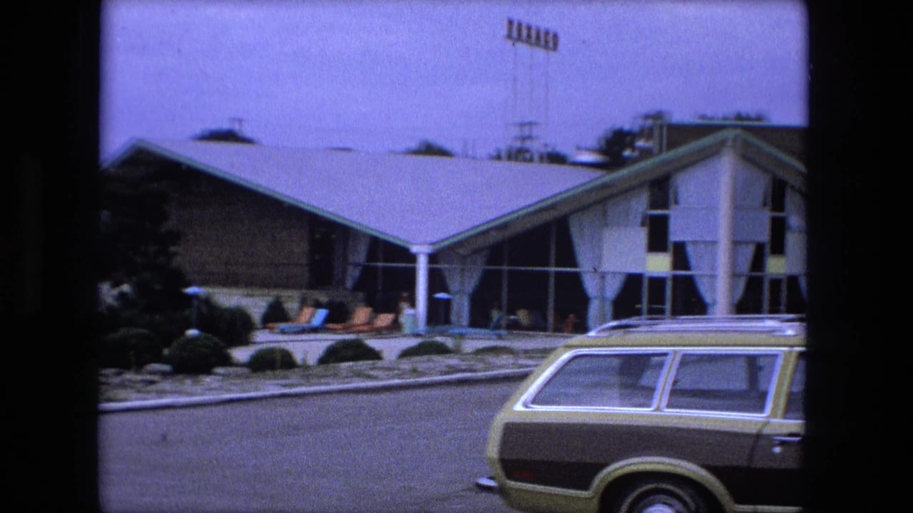 Station wagon passing a low modern motel or resort building with angled rooflines and patio seating.