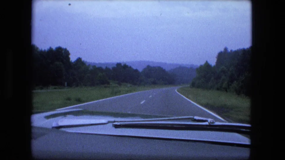 1974 home-movie driving view through a windshield on a curving rural Indiana highway under an overcast sky.