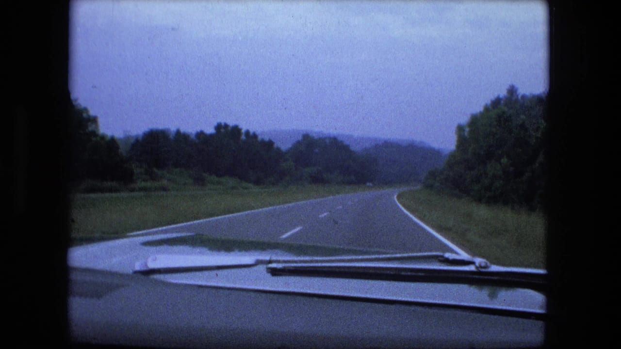 Forward view from a car on a curving two-lane highway bordered by trees and low hills.