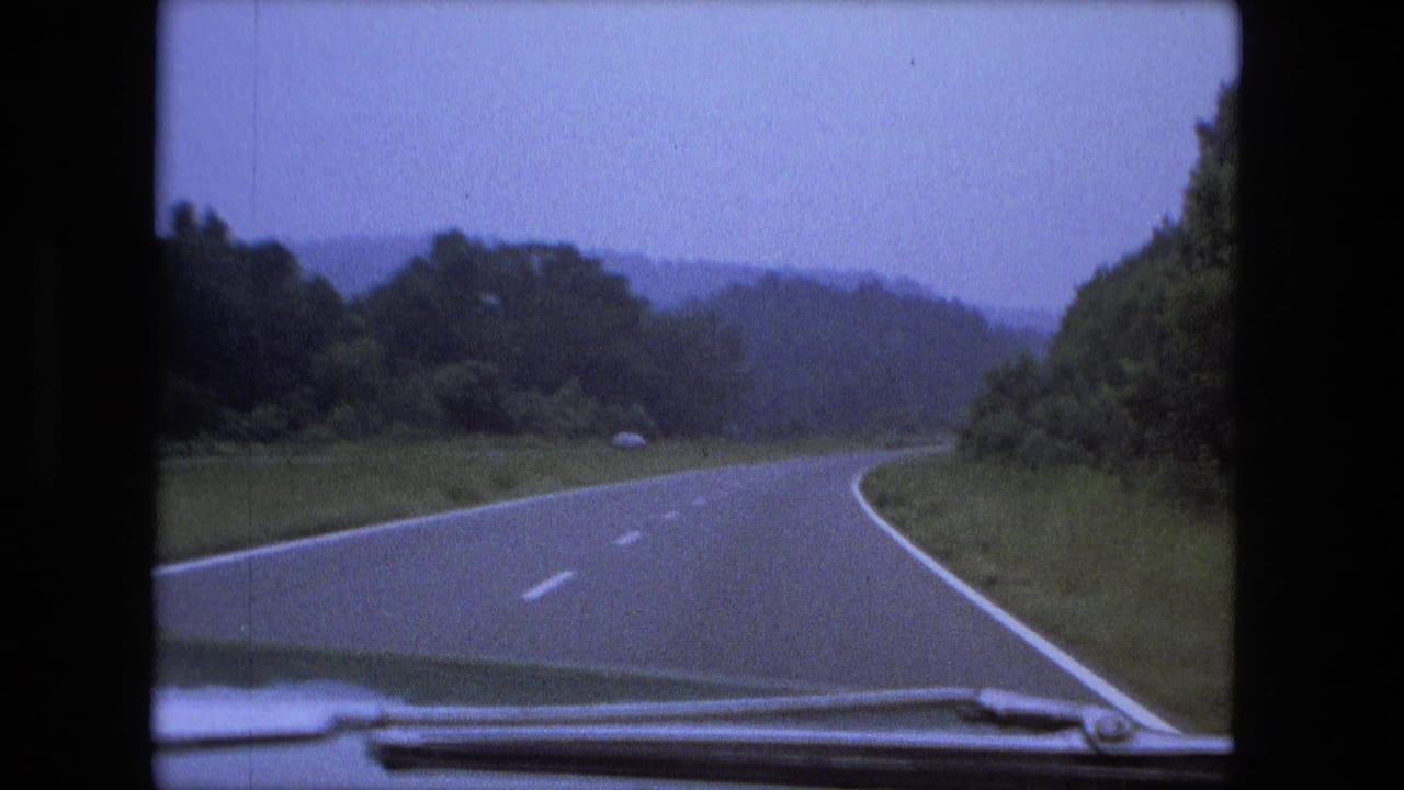 Windshield view of a right-curving rural highway in 1974 home-movie footage, with trees and distant hills under a muted sky.