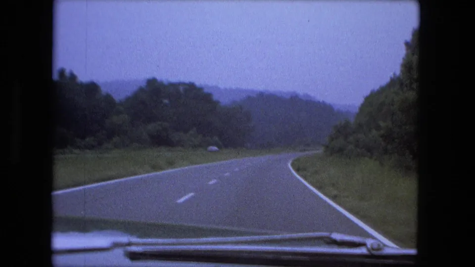 Windshield view of a right-curving rural highway in 1974 home-movie footage, with trees and distant hills under a muted sky.