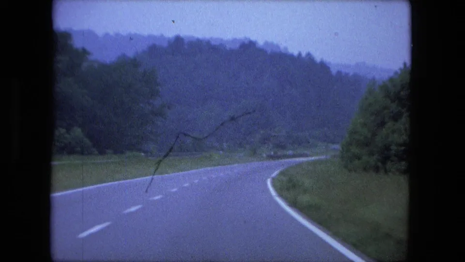 Soft 8mm view of a car following a curved wooded highway near Fort Wayne, with blue color shift and visible film marks.