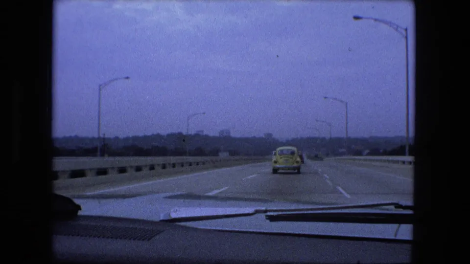 1974 windshield view on a bridge or divided roadway near Fort Wayne, with a yellow compact car ahead and streetlights along the road.