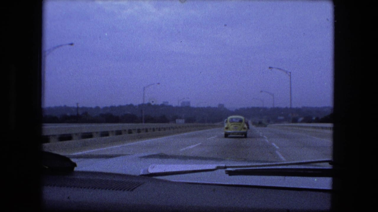 Windshield view of a highway with a yellow Volkswagen Beetle ahead under an overcast sky.