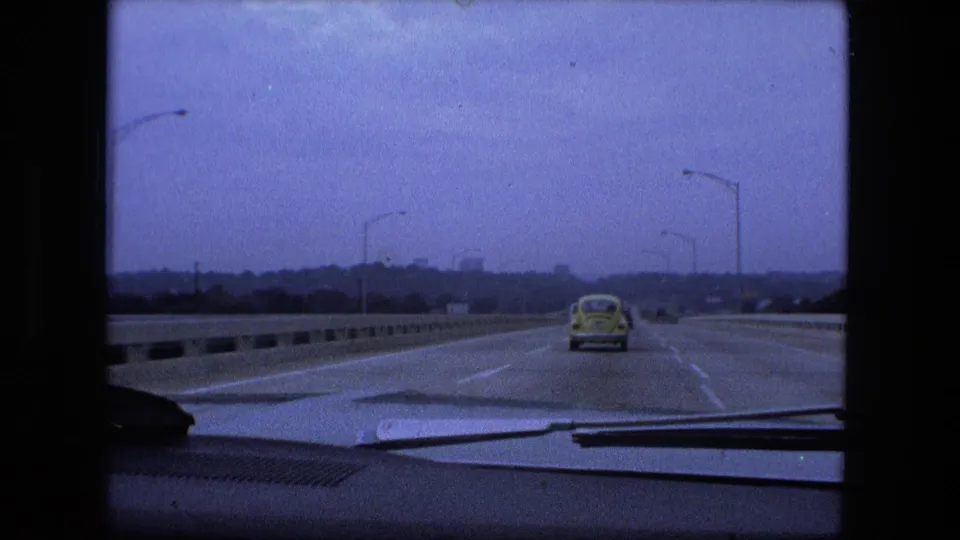 1974 Fort Wayne, Indiana 8mm frame showing a highway from inside a moving car, with a yellow Volkswagen Beetle ahead, concrete barriers, streetlights, and distant buildings.