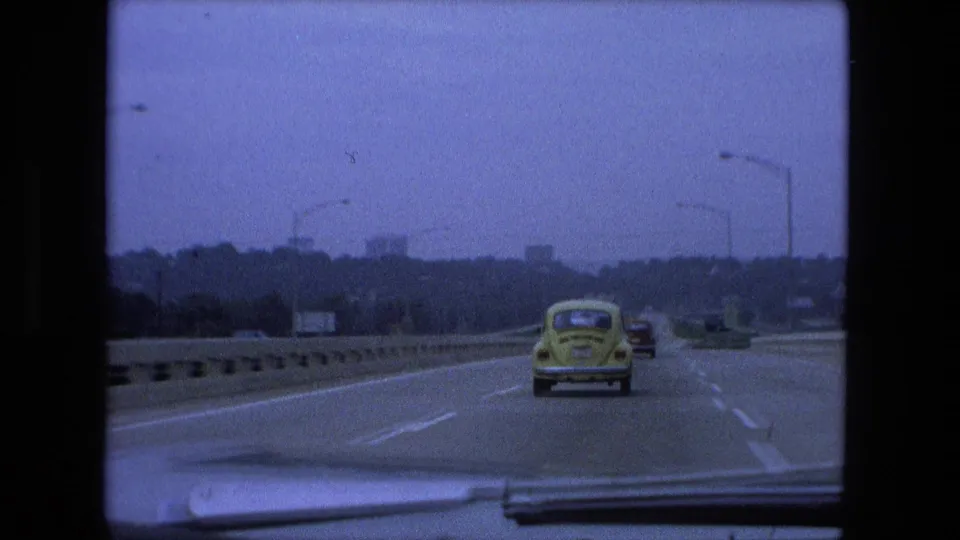 1974 Fort Wayne, Indiana highway view from a car, with the yellow Volkswagen Beetle centered ahead, other vehicles in the distance, roadside barriers, and tall streetlights.