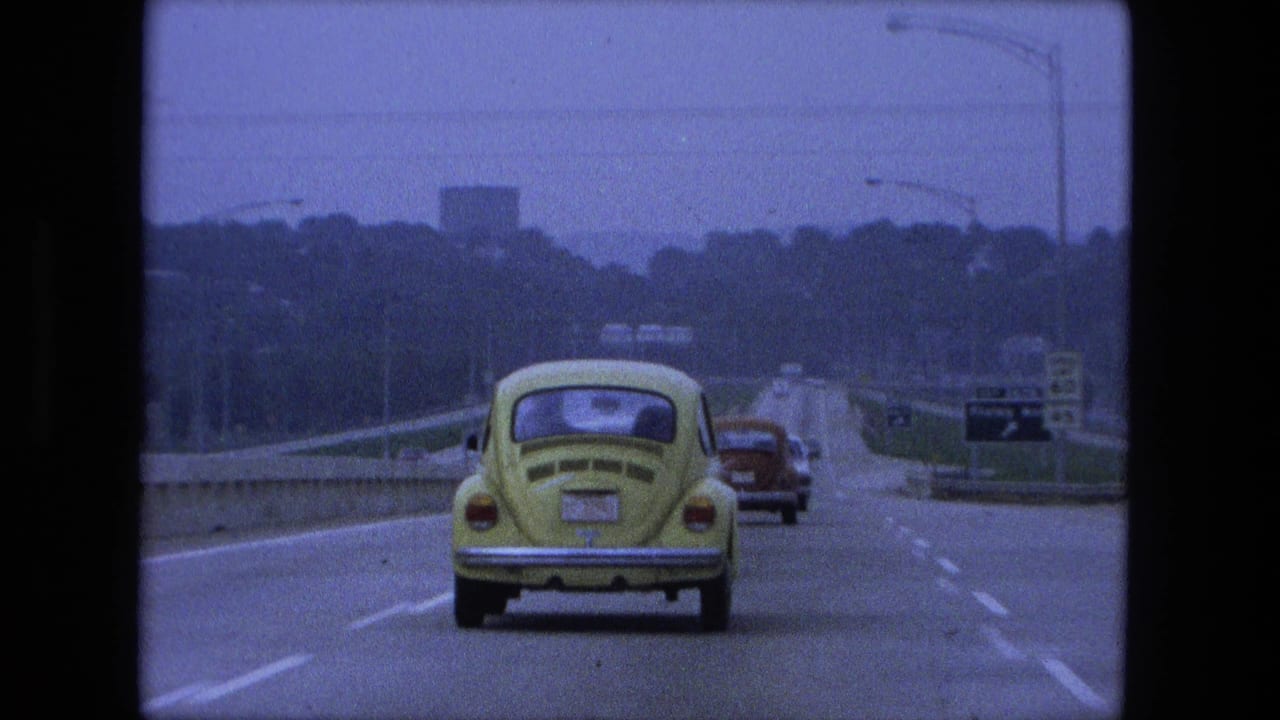 1974 Fort Wayne, Indiana frame from a moving car showing a closer view of the yellow Volkswagen Beetle, highway lanes, road signs, distant vehicles, and a tree-lined city approach.
