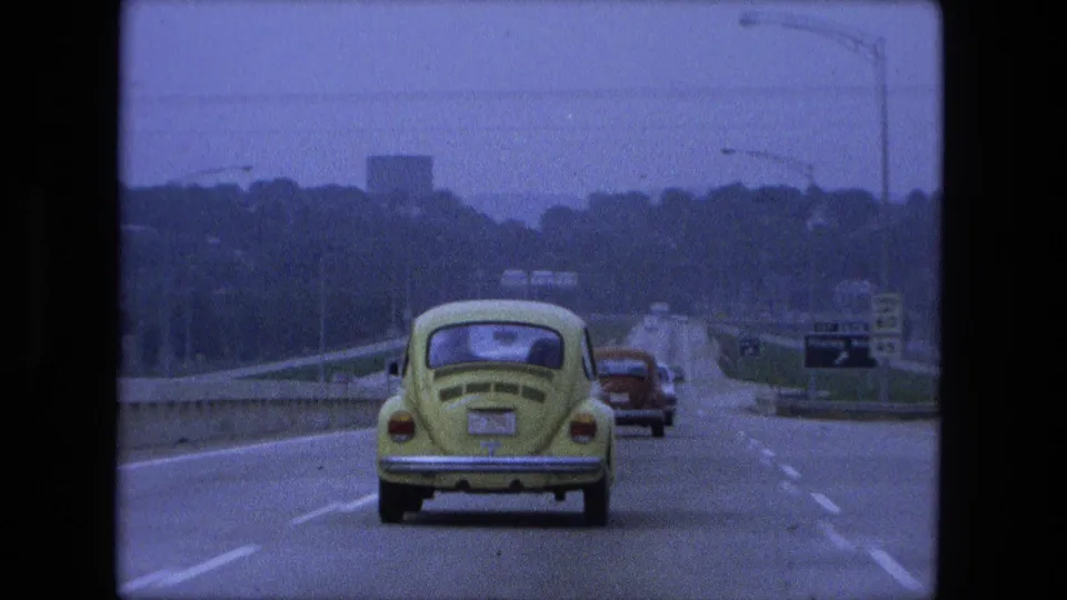 1974 Fort Wayne, Indiana frame from a moving car showing a closer view of the yellow Volkswagen Beetle, highway lanes, road signs, distant vehicles, and a tree-lined city approach.