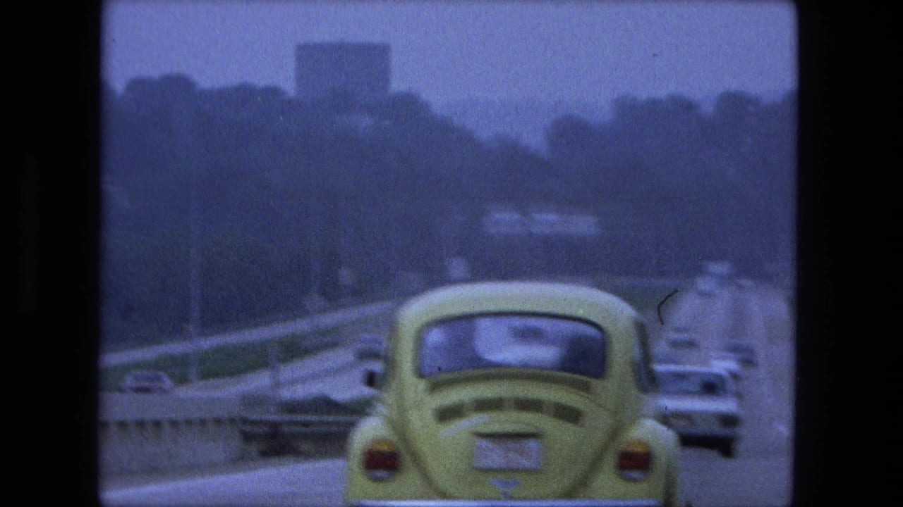 1974 Fort Wayne, Indiana 8mm frame with a close rear view of the yellow Volkswagen Beetle, a neighboring car, blurred roadside trees, highway barrier, and distant building shapes.