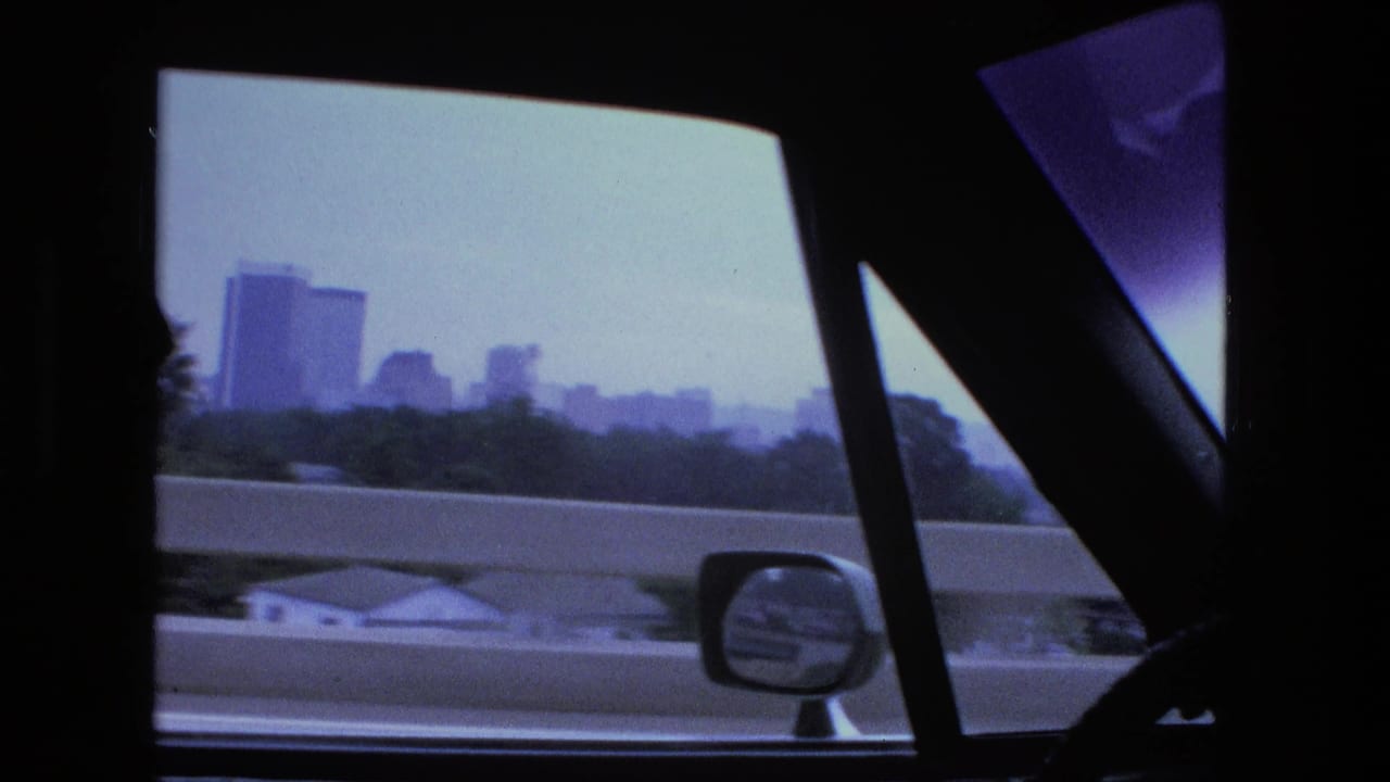 The moving car passes a highway barrier while a hazy Fort Wayne skyline, side mirror, and dark window frames fill the 1974 8mm frame.