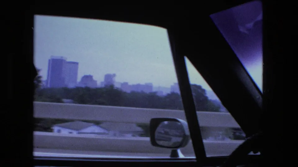 The moving car passes a highway barrier while a hazy Fort Wayne skyline, side mirror, and dark window frames fill the 1974 8mm frame.