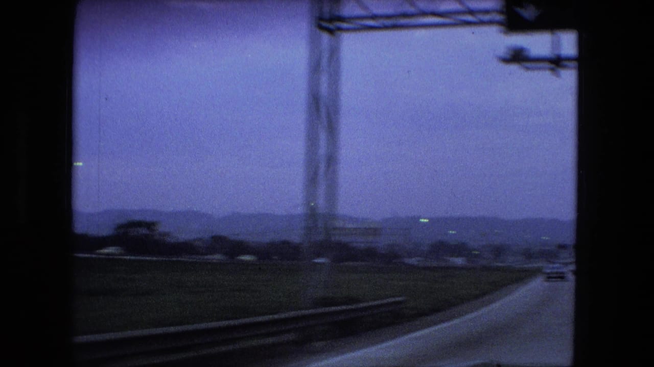 The car looks out toward a highway curve, roadside structure, low hills, and distant traffic under a blue-purple 8mm color cast in the Fort Wayne road-trip footage.