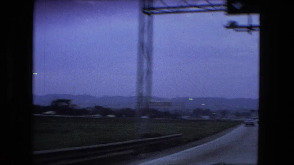 The car looks out toward a highway curve, roadside structure, low hills, and distant traffic under a blue-purple 8mm color cast in the Fort Wayne road-trip footage.