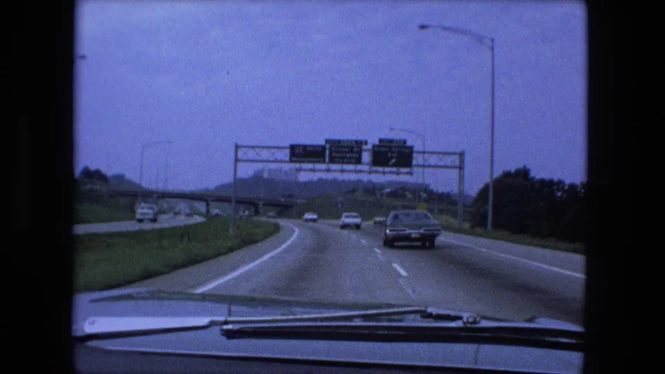 Windshield view of traffic approaching overhead highway signs near Fort Wayne, Indiana, with 1970s cars and roadside lights visible.