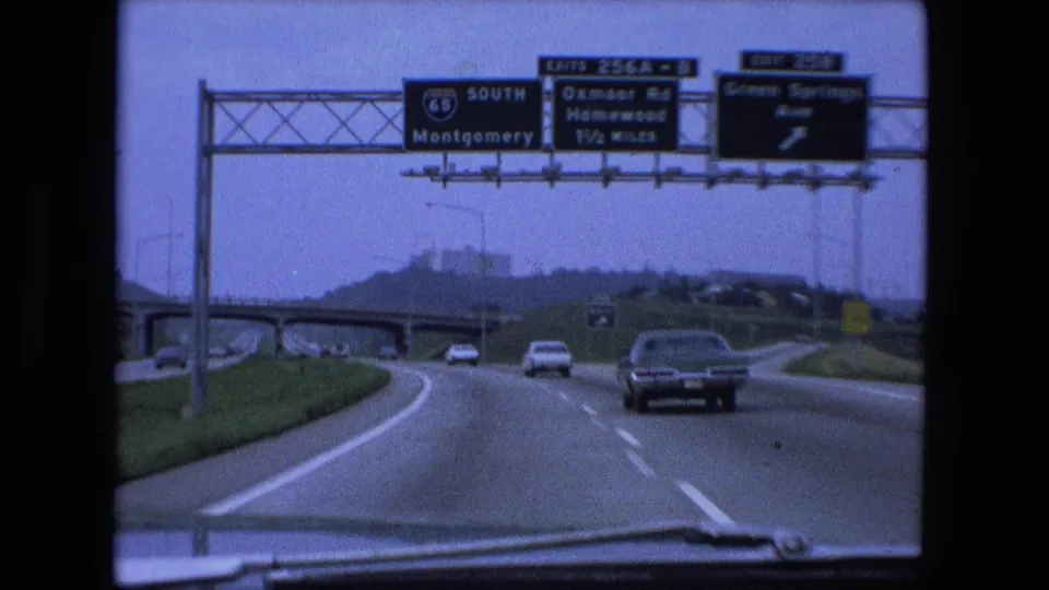 1974 passenger-view highway shot with overhead signs including I-65 South, moving cars, an overpass, and the car hood at the bottom edge.