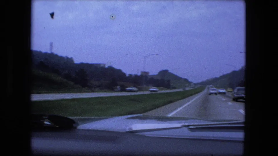 Dashboard-edge view from a moving car on a Fort Wayne interstate, with vehicles ahead, grassy median, hillsides, and roadside lights.