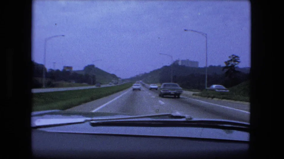 1974 Fort Wayne moving-car view showing interstate traffic, dashboard trim, road signs, streetlights, grassy embankments, and distant structures.
