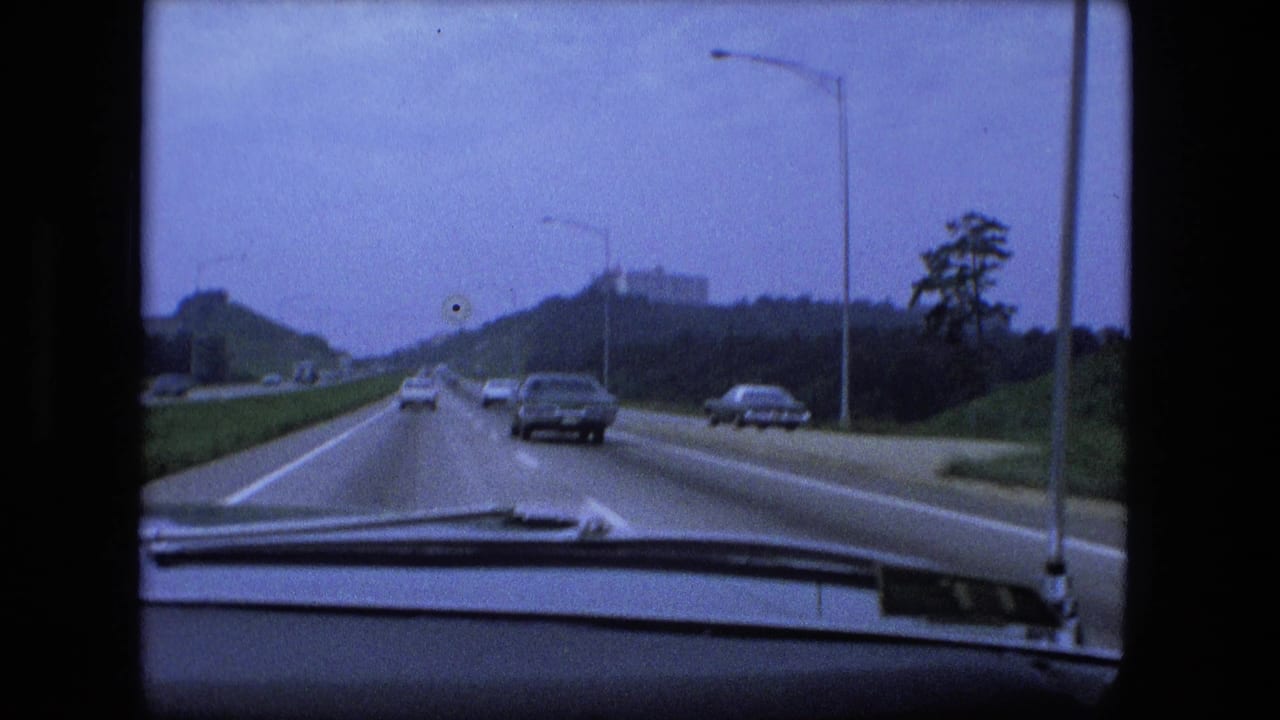 Forward view from a car on a multi-lane highway with cars ahead and a distant building on a hill.