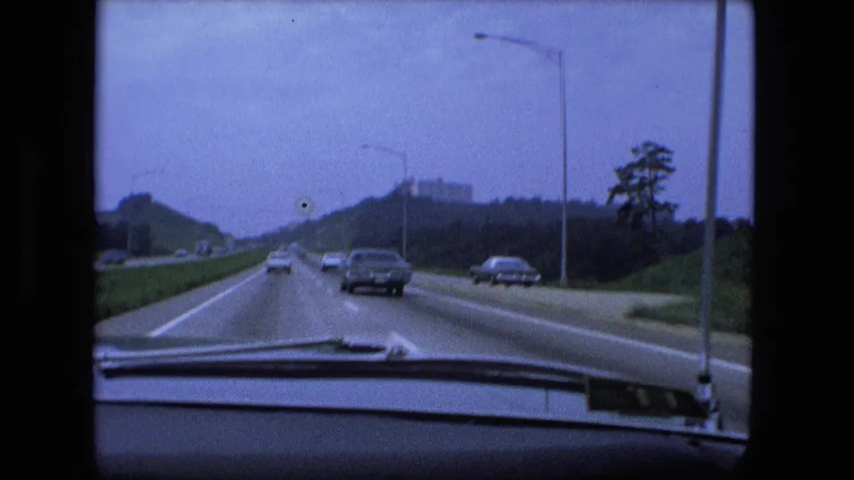 1974 Fort Wayne interstate view from inside a moving car, with period automobiles, highway lights, trees, and a distant hilltop building.