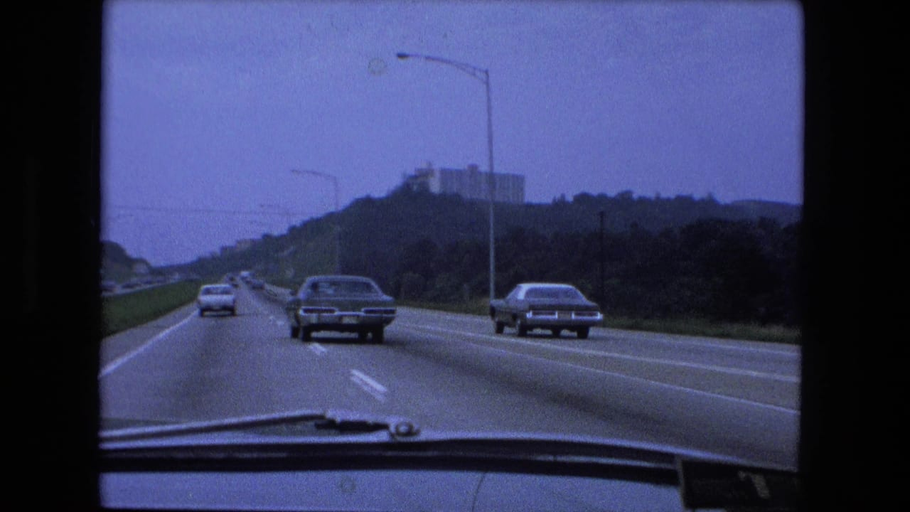 Windshield view of Fort Wayne-area highway traffic in 1974, showing classic cars, lane markings, roadside greenery, and a distant building.