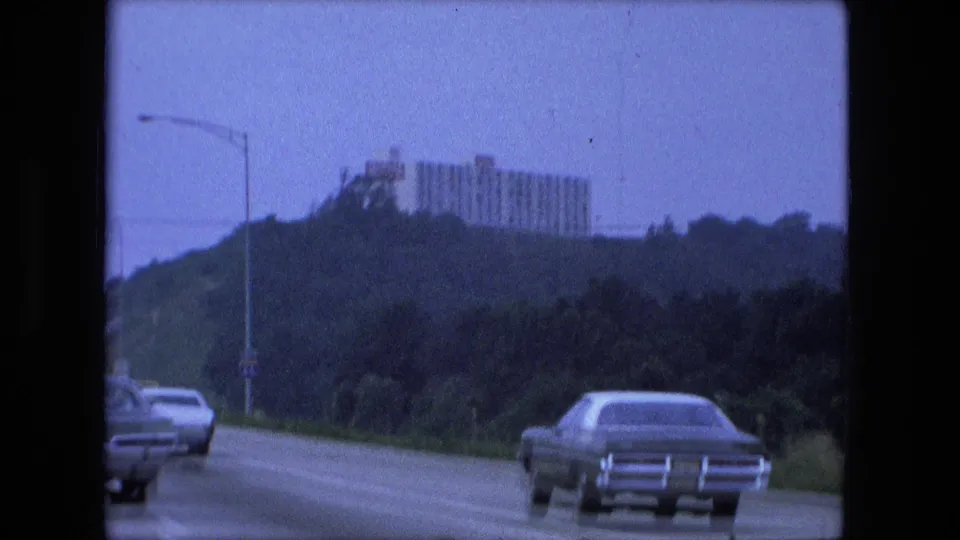 Fort Wayne highway scene from a moving car in 1974, with automobiles in the lanes and a large building rising above trees on a hill.