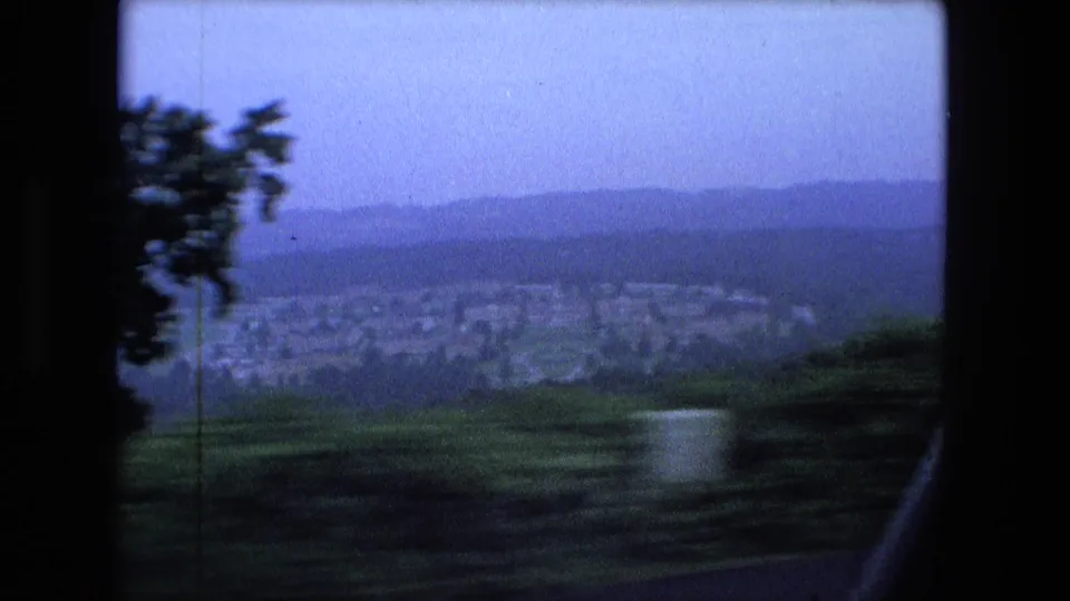 Moving-car view near Fort Wayne in 1974, with motion blur across roadside greenery and a distant built-up area under a hazy sky.