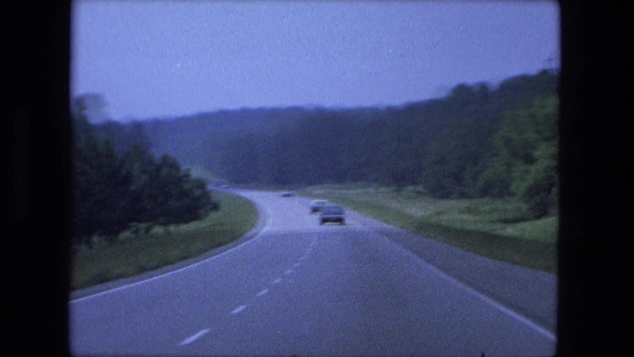Curving highway through wooded terrain with several cars ahead under a pale sky.