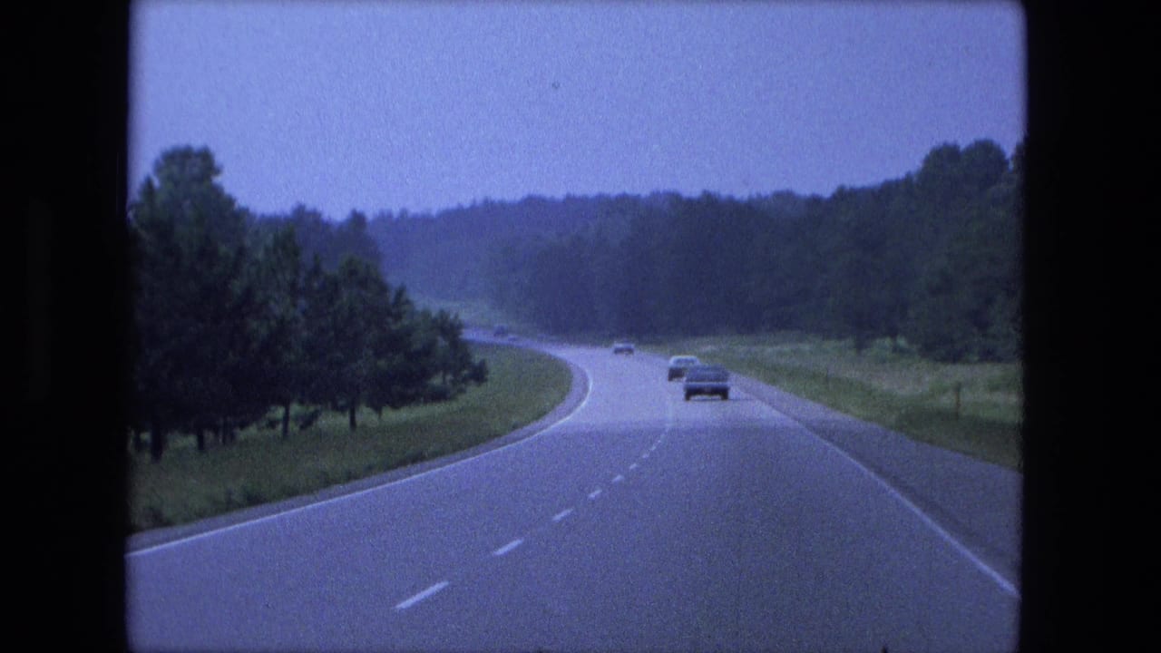 Wide highway lane curving left with cars ahead and trees lining both sides.
