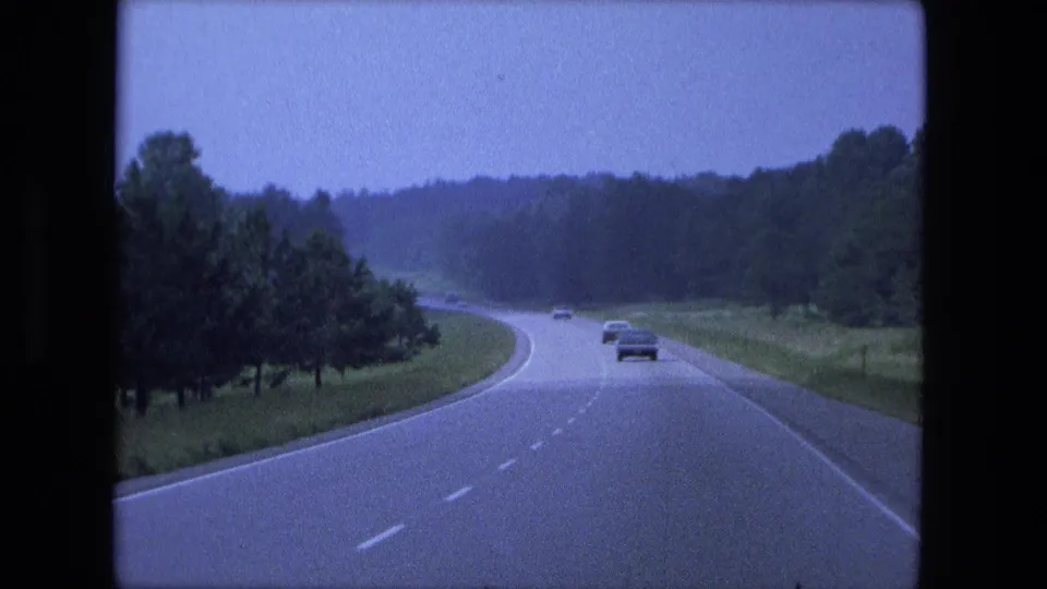 1974 Indiana highway driving view with cars ahead on a broad road, grassy shoulders, and dense trees in the distance.