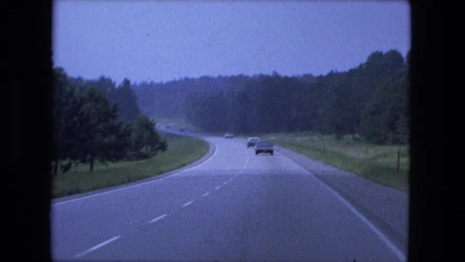 Home-movie frame from 1974 showing cars on a curving Fort Wayne area highway bordered by wooded and grassy land.