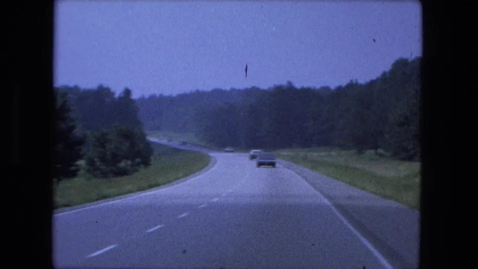 1974 8mm road-travel frame with cars receding along a wooded highway near Fort Wayne, Indiana.